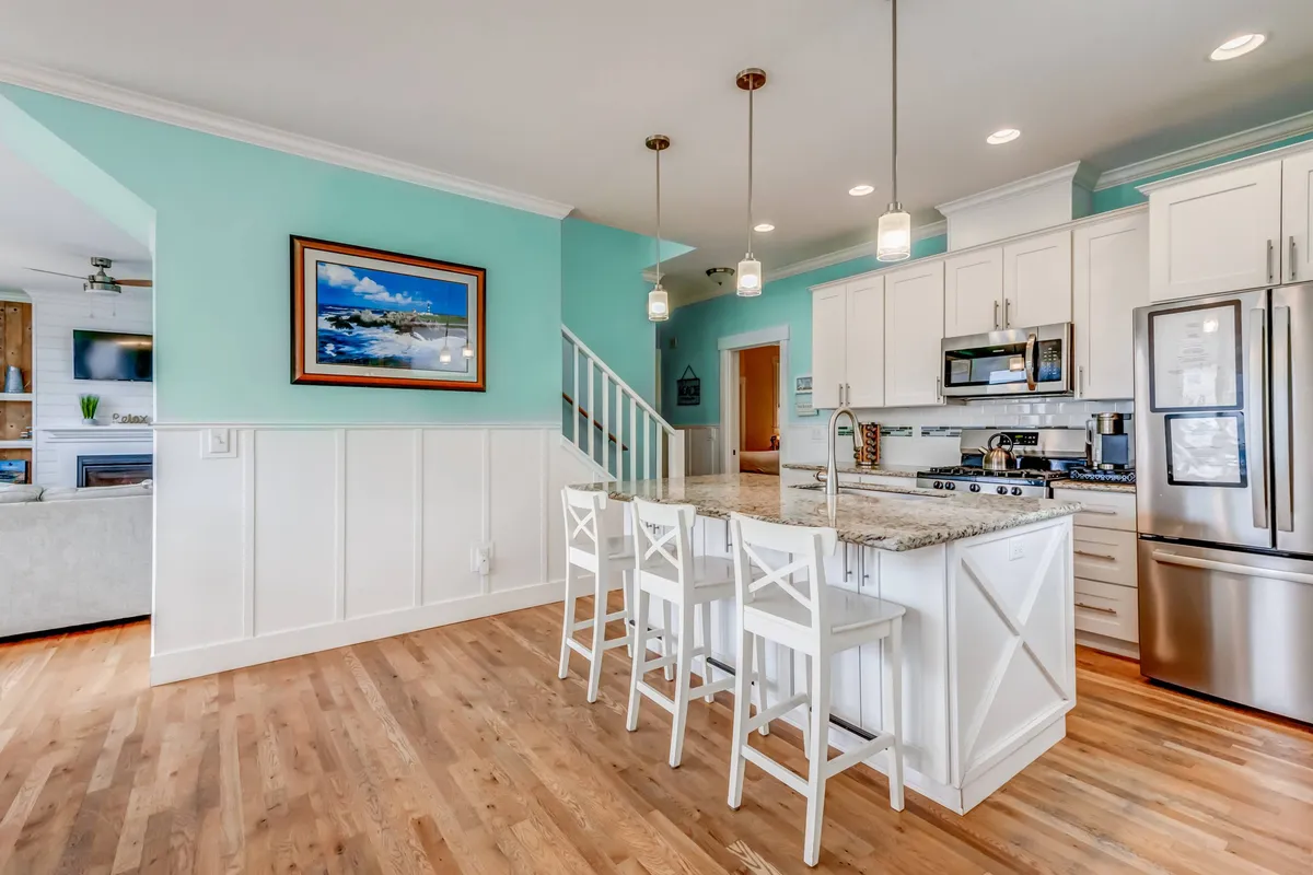 A bright, open kitchen featuring a granite island, white cabinetry, turquoise accent wall, and stylish pendant lights. The scene captures a cheerful moment of people cooking and enjoying each other's company in a welcoming, functional space.