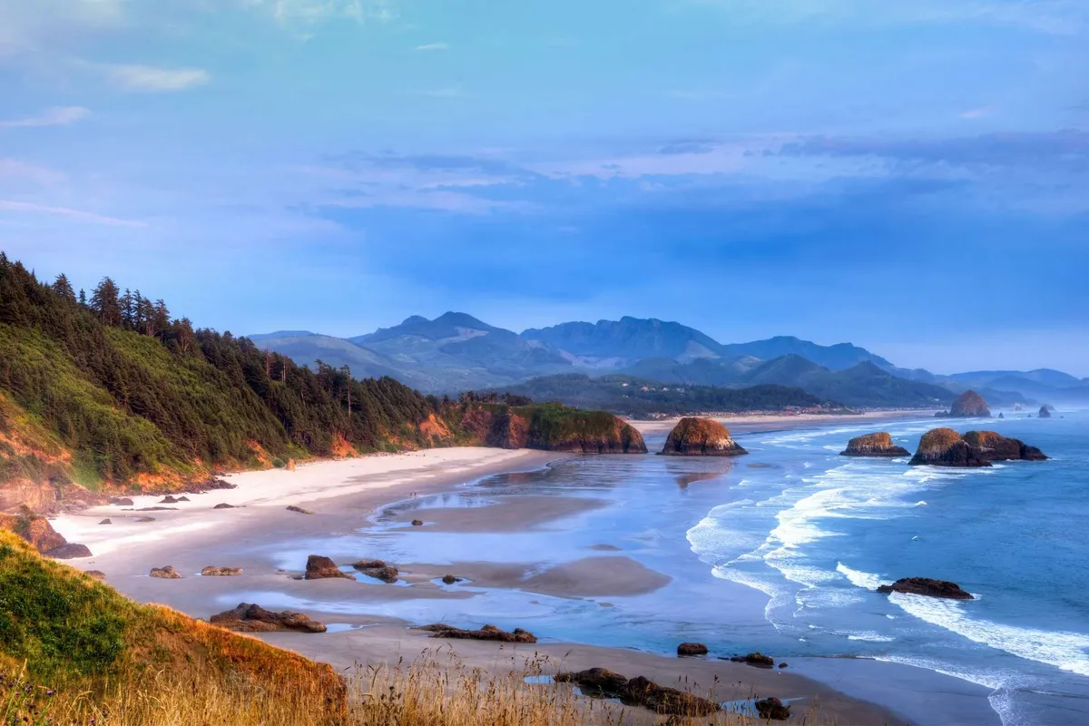 A stunning coastline view with ocean waves gently rolling onto a sandy beach, punctuated by rock formations and distant mountains under a serene sky. The scene captures the tranquil essence of the beach, inviting viewers to imagine sunset strolls, beachcombing, and moments of relaxation by the water.