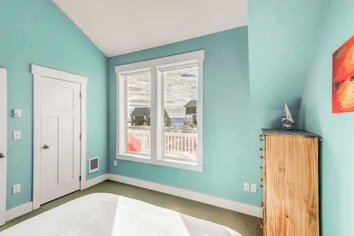 This interior scene features large windows framing what appears to be an enticing outdoor view, with bright blue walls and wooden furniture creating an inviting, airy atmosphere. The focus is on the connection between the comfortable indoor space and the natural world outside, hinting at the coastal scenery just beyond the windows.