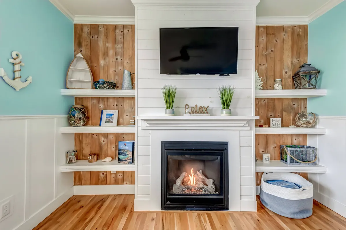 A tranquil living space with a modern fireplace, wall-mounted television, and open shelving adorned with books and coastal-themed decor. The room feels both comfortable and stylish, ideal for relaxing or catching up on shows.