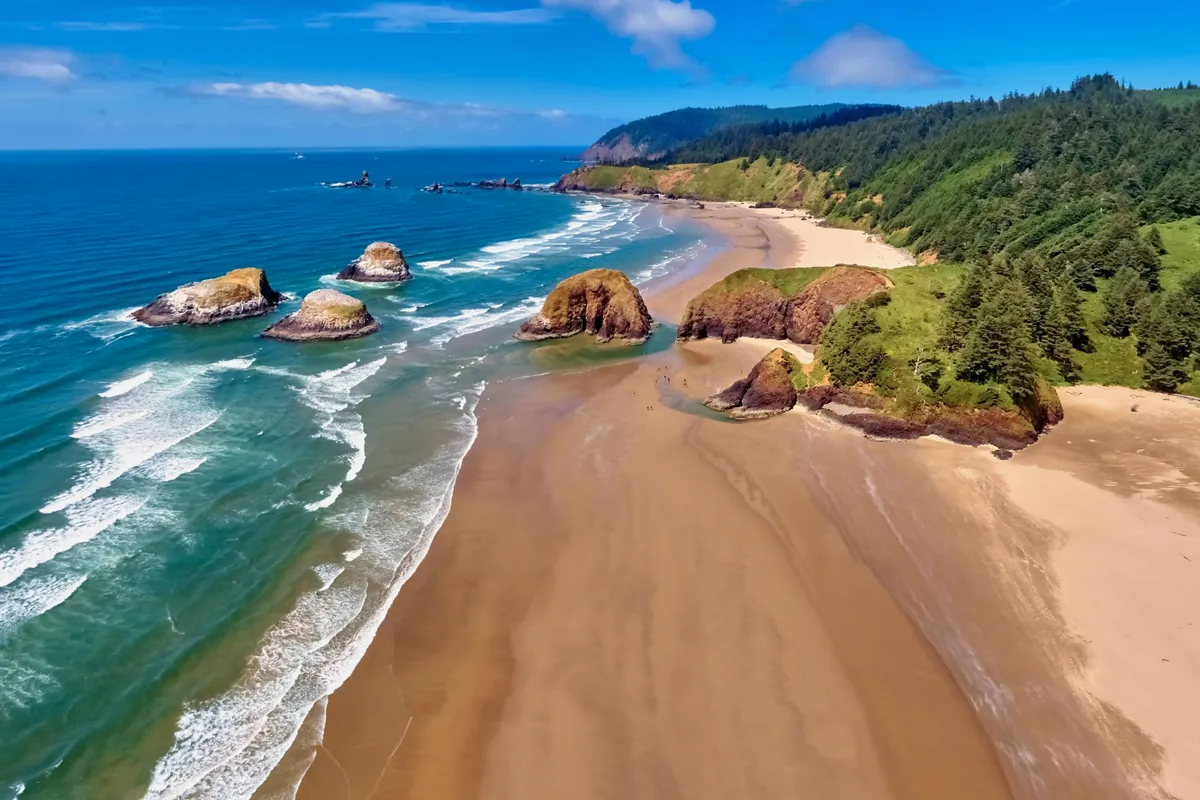 A wide shot of a sandy beach bordered by lush greenery and striking rock formations, with waves crashing onto the shore. The vibrant coastal environment invites exploration and embodies the outdoor amenities and activities available to guests.