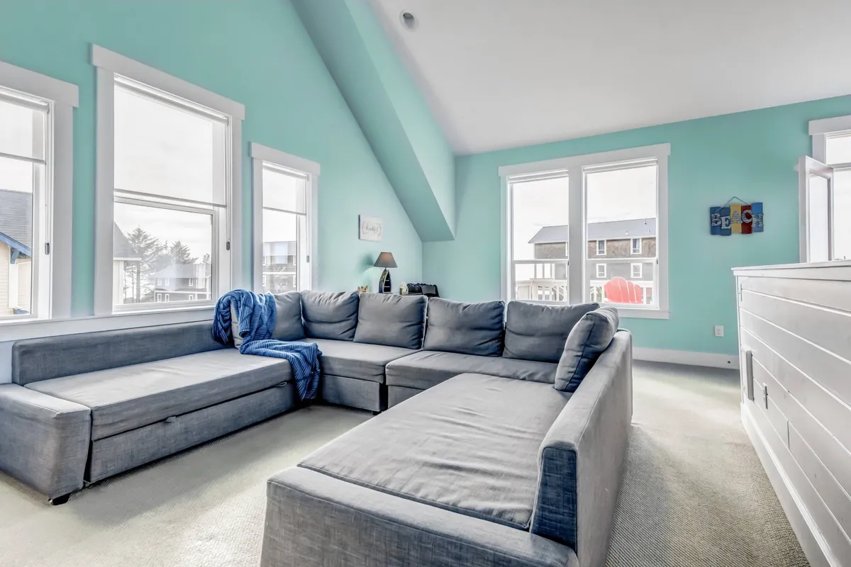 A spacious, inviting media/game room with a large sectional sofa, colorful wall art, and windows letting in plenty of natural light. The setting suggests a flexible, multi-use space ideal for group gatherings, movie nights, or extra sleeping arrangements.
