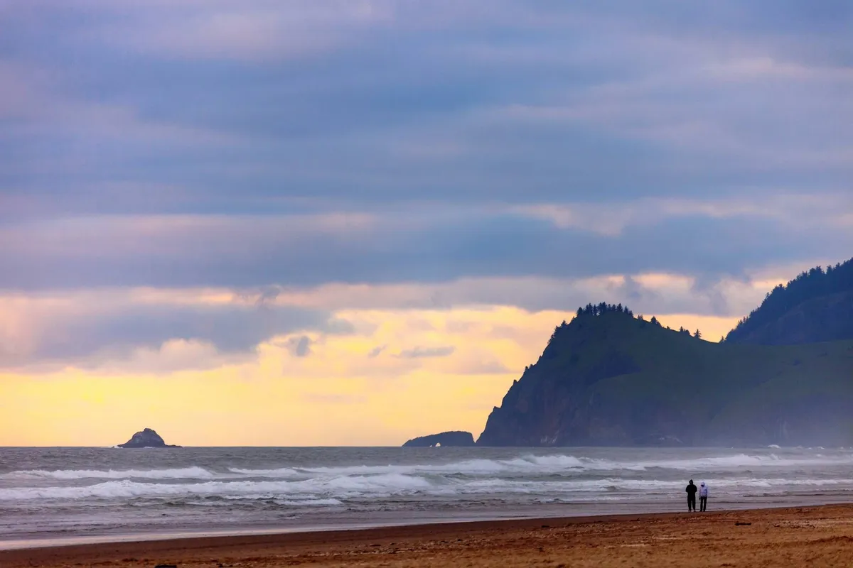 A sweeping view of the sandy beach with gentle ocean waves rolling in and distant cliffs on the horizon. Two figures walk together along the shoreline, emphasizing the accessible and family-friendly nature of the coast. The lighting hints at an inviting sunset, perfect for creating memories during strolls or beachcombing adventures.