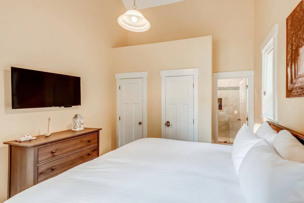 A relaxing queen bedroom with a plush bed dressed in white linens, a dresser, a television, tasteful wall art, and a door leading to an en suite or Jack & Jill bath. The space feels cozy and functional, perfect for couples or guests seeking privacy and convenience.