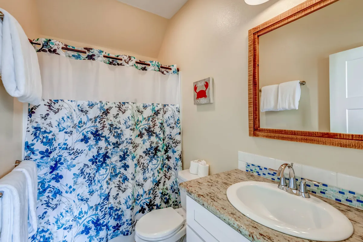 A light-filled bathroom featuring a floral shower curtain, granite countertops, a large mirror, and whimsical crab artwork. The space exudes a sense of tranquility, offering an inviting spot for refreshing showers or soothing evening routines.