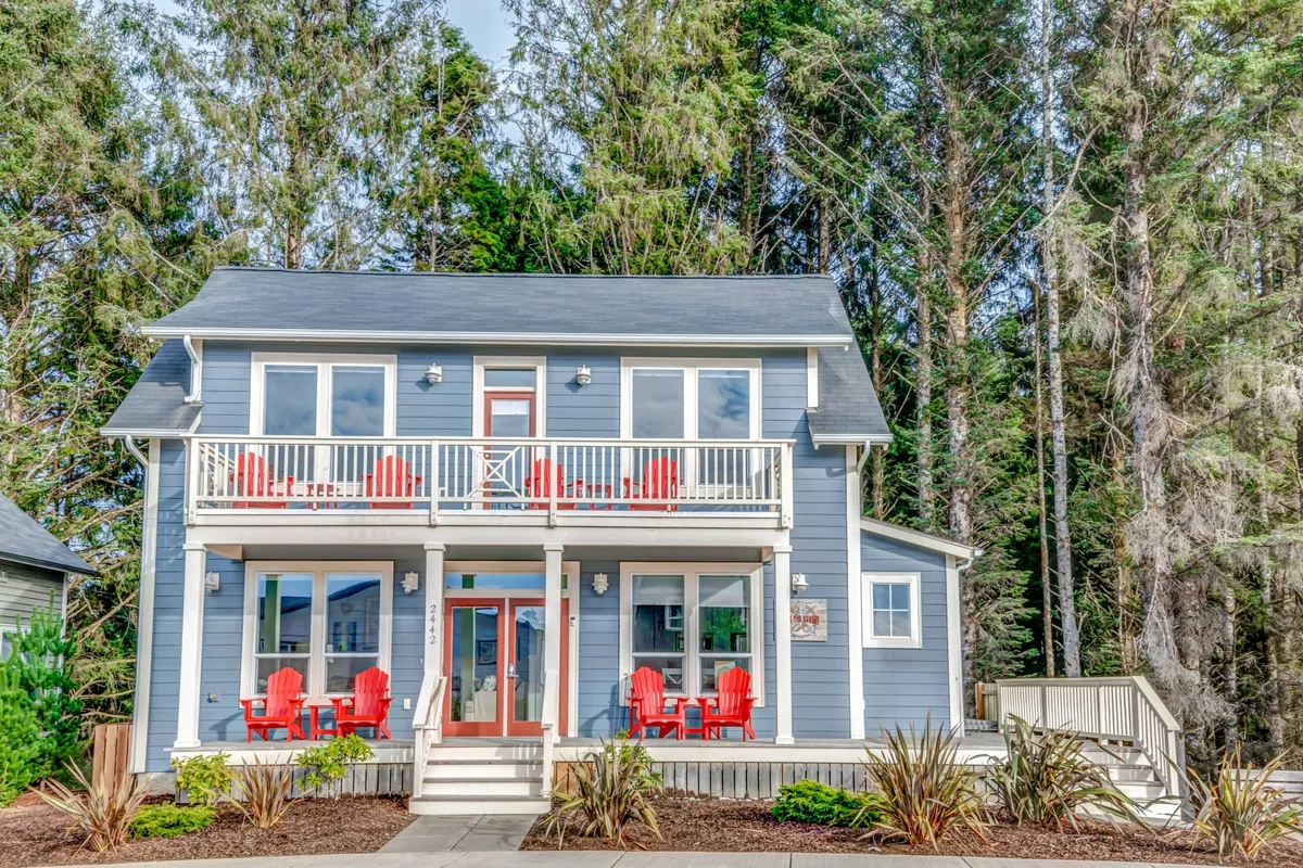 The hero image showcases the home's charming exterior, featuring a welcoming front porch adorned with inviting red chairs, expansive windows that hint at airy interiors, lush green trees surrounding the house, and a tranquil, sunset-lit atmosphere perfect for relaxing or gathering with family and friends.