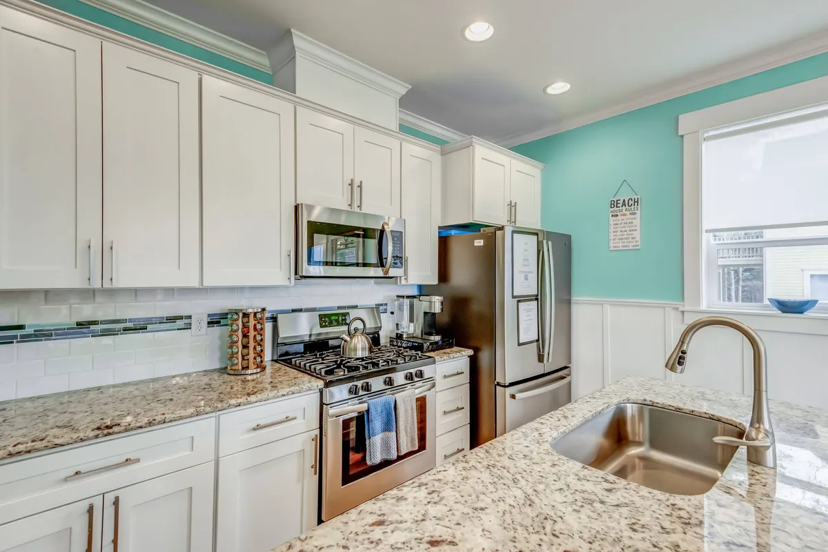 A bright, inviting kitchen with granite countertops, stainless steel appliances, and playful beach-themed decor set against a vibrant turquoise wall. The space feels open and lively, ideal for gathering with friends or family after a day at the coast.