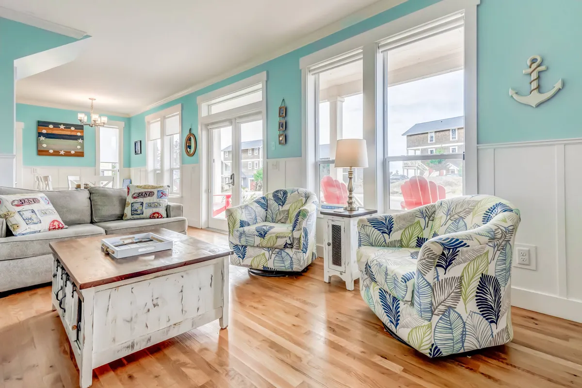 A bright and vibrant living area featuring a comfortable sofa, colorful accent chairs, a large coffee table, and expansive windows framing sunrise views. The space is designed for socializing, relaxing, and enjoying time with friends and family.