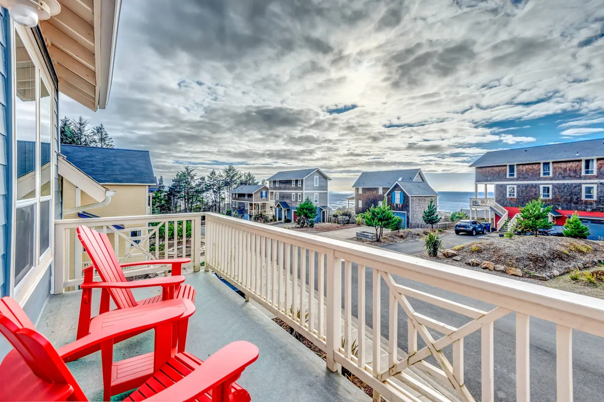 A cheerful exterior deck scene with red Adirondack chairs facing out toward an ocean view, flanked by neighboring houses. The inviting setup suggests a perfect spot for sunset watching, relaxing, and socializing with family or friends.