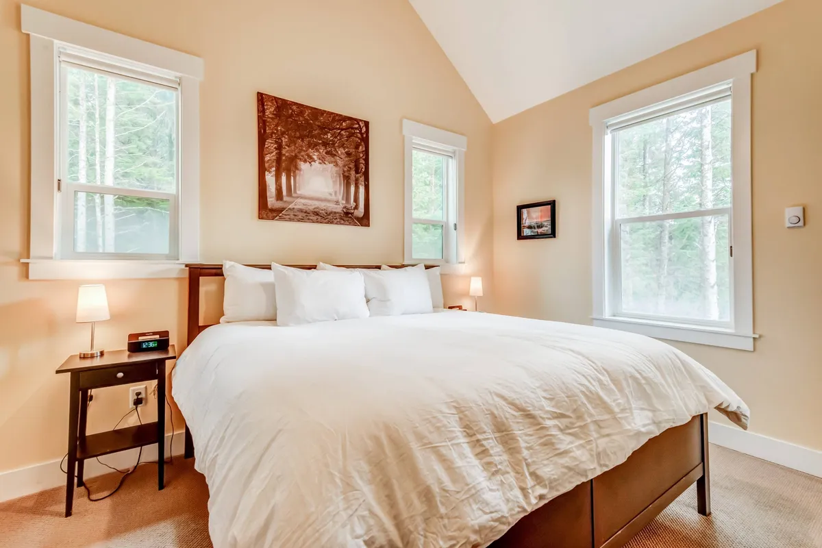A spacious bedroom featuring a king-sized bed with crisp white linens, tasteful artwork, matching nightstands with lamps, and large windows that let in abundant natural light, suggesting views of the surrounding forest. The room exudes tranquility and offers plenty of space and comfort, ideal for a restful retreat.